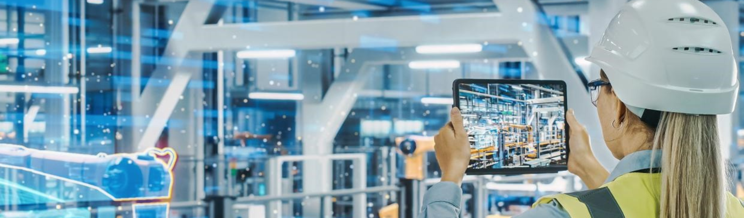 A person wearing a safety helmet uses a tablet to monitor a modern, automated factory with robots and digital visualizations.