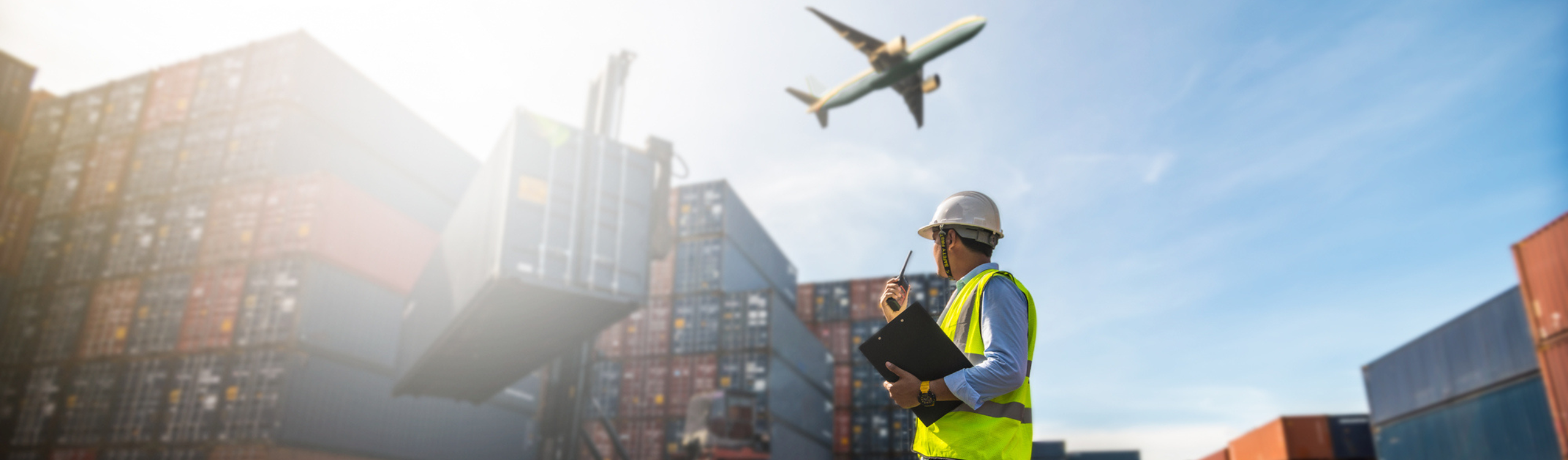 Man standing in front of stacked shipping containers, holding a clipboard and using a pager, while containers are being loaded in the background and a plane is taking off.