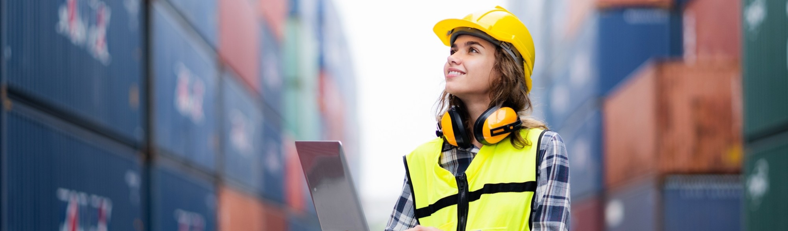A woman wearing a warning vest and a safety helmet works on a laptop at the container port, surrounded by shipping containers – international goods exports, logistics, and trade with India