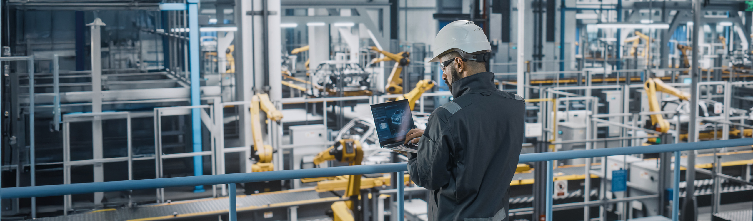 A worker wearing a safety helmet and protective clothing stands on an elevated platform in a modern industrial facility, operating a laptop. In the background, several yellow industrial robotic arms and automated production systems are visible.
